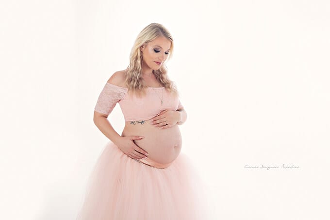 Blonde schwangere Frau im rosa Rock und in der Bluse, die ihren Bauch waehrend des Fotoshootings in Carmen Bergmann-Fotostudio in Haidhausen Muenchen schaut