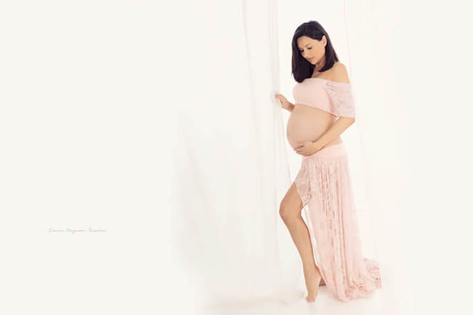 Schwangere Frau mit langem dunklem Haar und rosafarbenem Kleid die neben einem Fenster steht und ihren Bauch im Carmen Bergmann-Fotostudio in Muenchen beobachtet